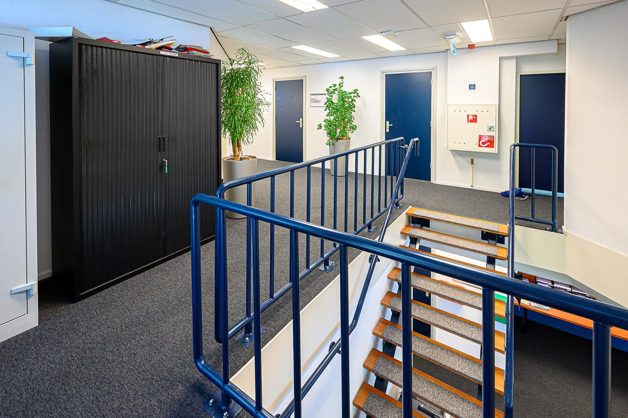 Trapportaal in een kantoorgebouw met blauwe deuren, planten en archiefkasten.